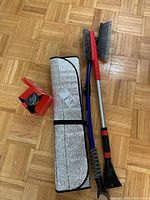 Photo showing the reflective car window shade folded and secured with a strap, the car adapter battery charger, and two snow brushes with their handles and scrapers visible on a parquet floor.