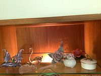 Display of assorted glass and porcelain decorative items including swan and butterfly figurines, flower ornaments, and lidded boxes on a wood shelf with backlight