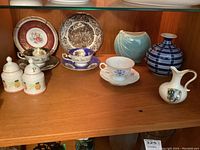 Photo showing nine China pieces arranged on a wooden shelf, including cups, saucers, mini plates, salt and pepper shakers, and decorative vases.