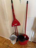 Photo showing a red Vileda Oscar broom and matching dustpan with long handles, mop with handle inside a red mop bucket with black wringer, and two plastic bins (one with flower pattern, the other smaller and grey). Also shows an extra red pole standalone.