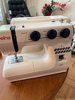 Full view of Elna HD22 sewing machine showing the white body with red 'elna' on the side and various control knobs for stitch selection, length, and width settings.