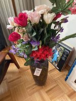 Full view of multi-colored faux floral arrangement in mottled dark stone vase on floor next to a chair and some other items.