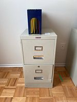 Front view of beige metal filing cabinet with two drawers and metal label holders, with blue file folder organizer containing yellow and red folders on top.