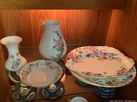 Five floral china pieces displayed in a wood display case with soft lighting: includes vases and plates with multi-color floral patterns.