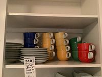 Shelf holding stacks of colorful Harmony Block cups and white saucers, total 18 cups and 18 saucers visible.