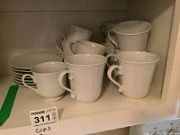 Shelf with six white IKEA china cups and saucers, plus additional mixed white cups and saucers. Cups vary in style; some cylindrical with handles, some smaller with flared lips. Saucer styles also vary. All in worn condition with some discoloration.