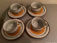 Four two-piece soup cups and matching saucers arranged in a 2x2 pattern on a flat surface, showing the orange and brown banded designs with speckled white ceramic.