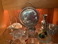 Photo showing the metal and glass decorative items arranged on a wooden surface with warm lighting. Items include a large metal plate with handle, metal vases, Dansk bowl, and various clear glass dishes and decorative glass ball.
