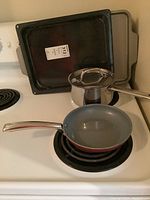 Full view of stovetop with small green frying pan on burner, stainless steel IKEA saucepan with lid behind it, and two metal baking trays stacked upright behind saucepan