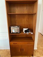 Front view of tall teak veneer cabinet showing shelves with some items, a drawer with handles, and closed cabinet doors below.