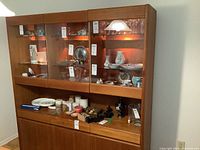 Front and right side view of teak vintage cabinet with three illuminated glass display cabinets containing various decorative items, three drawers below the display section, and three lower storage compartments with closed doors.