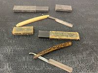 Top view of two antique straight razors lying on a black textured surface with their cases. The upper razor has a yellow handle and metal blade marked 'THE-MAB'. The lower razor has a faux tiger-striped handle with a curved frame and a matching mottled case.