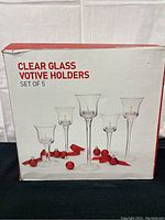 Front of original box showing set of 5 clear glass votive holders with tapered, flared rims and varying stem heights.
