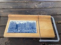 Front view of wooden cheese cutting board with blue Holland Delft tile featuring a cityscape and attached wire slicer.