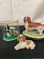 Lot of three dog figurines shown on black cloth: a large standing dog on green base, a lying dog in front, and a case with three smaller black and white dogs inside.
