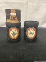 Two black tire-shaped drink holders with labels saying 'No.1 Grandpa and all round great guy'. One with packaging card reading 'Drink Holder'.