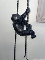 Close-up of single climbing man sculpture gripping twisted metal rod rope, showing figure's form and bronze texture.