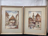 Two framed watercolor paintings displayed side by side on a white fabric background showing old European village scenes.