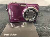 Photo of the purple Kodak EasyShare C195 digital camera front showing lens and branding, resting on top of a black user guide.