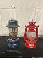 Front view of both lanterns showing the blue Coleman Deluxe Gas-Lite Lantern on left and red kerosene lantern on right.