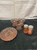 Pink depression glassware set including a saucer, three dessert bowls, and two salt and pepper shakers arranged on a black surface.
