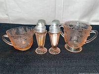 Full set of pink depression glass items: creamer, sugar dish, salt and pepper shakers on black fabric background.