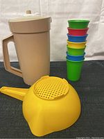 Photo showing beige Tupperware jug with lid, 6 colorful cups stacked, and yellow plastic strainer