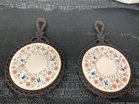 Pair of vintage cast iron pot rests with round ceramic inlays showing floral and duck motifs on a dark textured background.