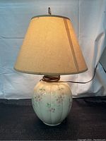 Full view of the vintage table lamp lit showing beige fabric shade and ribbed ceramic base with floral design.