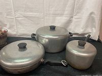 Three vintage cast aluminum cookware items: two pots with lids and one frying pan with lid, all with black handles, arranged against white background.