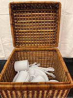 Open wicker picnic basket with white ceramic dishware inside.