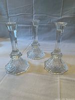 Three clear crystal candlestick holders, hexagonal bases with diamond cut pattern, flared top rim, standing on white cloth background against neutral backdrop.
