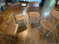 Four brown leatherette folding chairs arranged on hardwood floor. Metal frames show some scratches and use marks.