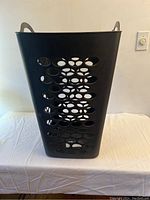 Black flexible square laundry hamper with oval ventilation holes, two carrying handles, placed on a white surface against a wall.