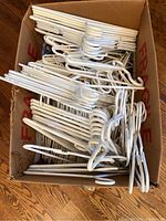 Box filled with numerous white plastic hangers, adult size, showing quantity and condition