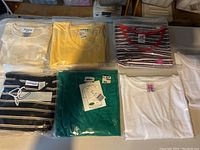 Six short sleeve ladies blouses folded on a table, showing multiple colors and patterns including solid white, yellow, teal, black-and-white stripes, and a red-trimmed striped blouse, some with original tags.