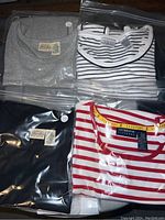 Four sleeveless tops in plastic bags showing grey, black, striped white and black, and red and white striped tops with tags.