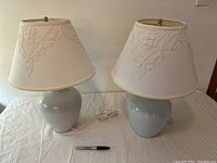 Pair of light grey-blue ceramic table lamps with white fabric shades featuring lace-type embroidery placed on a white surface with a sharpie marker for scale.