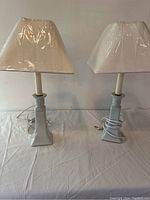 Pair of two light grey/blue ceramic candlestick-style table lamps with white shades, wrapped in plastic. Lamps are on a white cloth surface against a white wall.