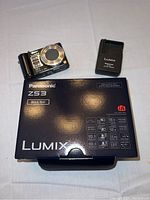 Photo showing Panasonic Lumix ZS3 camera, battery charger, and original product box on a flat surface.