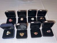 Full overview of 8 costume rings displayed in black individual boxes, showing various colorful stones in gold-plated bands, arranged in two rows.