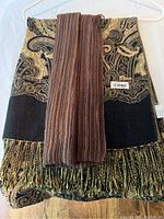 Black and gold paisley patterned scarf with fringes and brown striped scarf overlaid on a hanger against white background