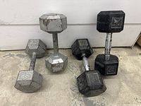 Photo showing two gray steel hex dumbbells and two black rubber coated hex dumbbells with chrome handles arranged against a white wall on a concrete floor.
