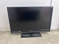 Front view of black Sharp Aquos flat screen TV showing the screen and base stand on a dusty floor against a plain wall.