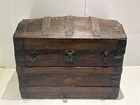 Front view of antique wooden humpback trunk showing metal lock hasp, latch closures, wood panels, and metal straps over wooden curved lid.