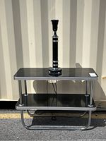 Front view of two-tier black laminate table with chrome frame and accompanying lamp mounted on top shelf