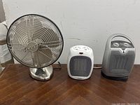 Front view of metal desk fan and two portable heaters on a wooden surface, showing their design and controls.