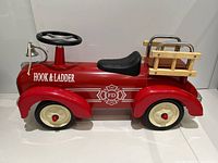 Side view of red Hook & Ladder kids ride-on firetruck showing metal body, black seat, cream plastic wheels, and wooden ladder rack.