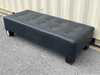 Angled side view of the long black leather buttoned ottoman on an outdoor surface, showing footprint and button tufting.
