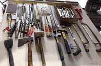 Assorted hand tools laid out on a table including hammers, saws, chisels, files, and wrenches spread in rows